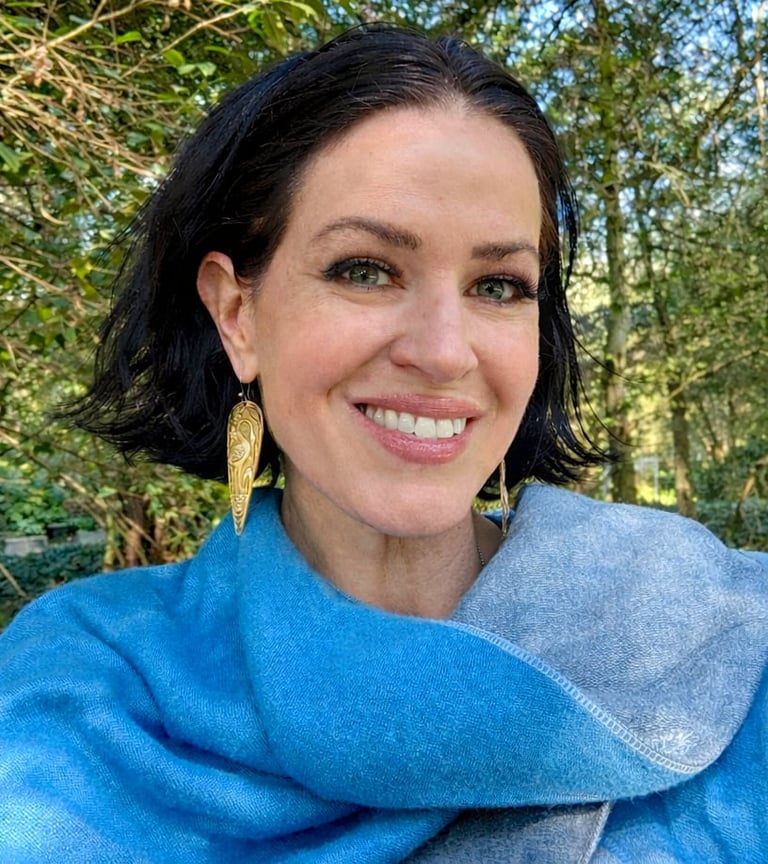 Woman with dark hair smiling at camera wearing blue sweater with gold earrings, outdoor greenery background